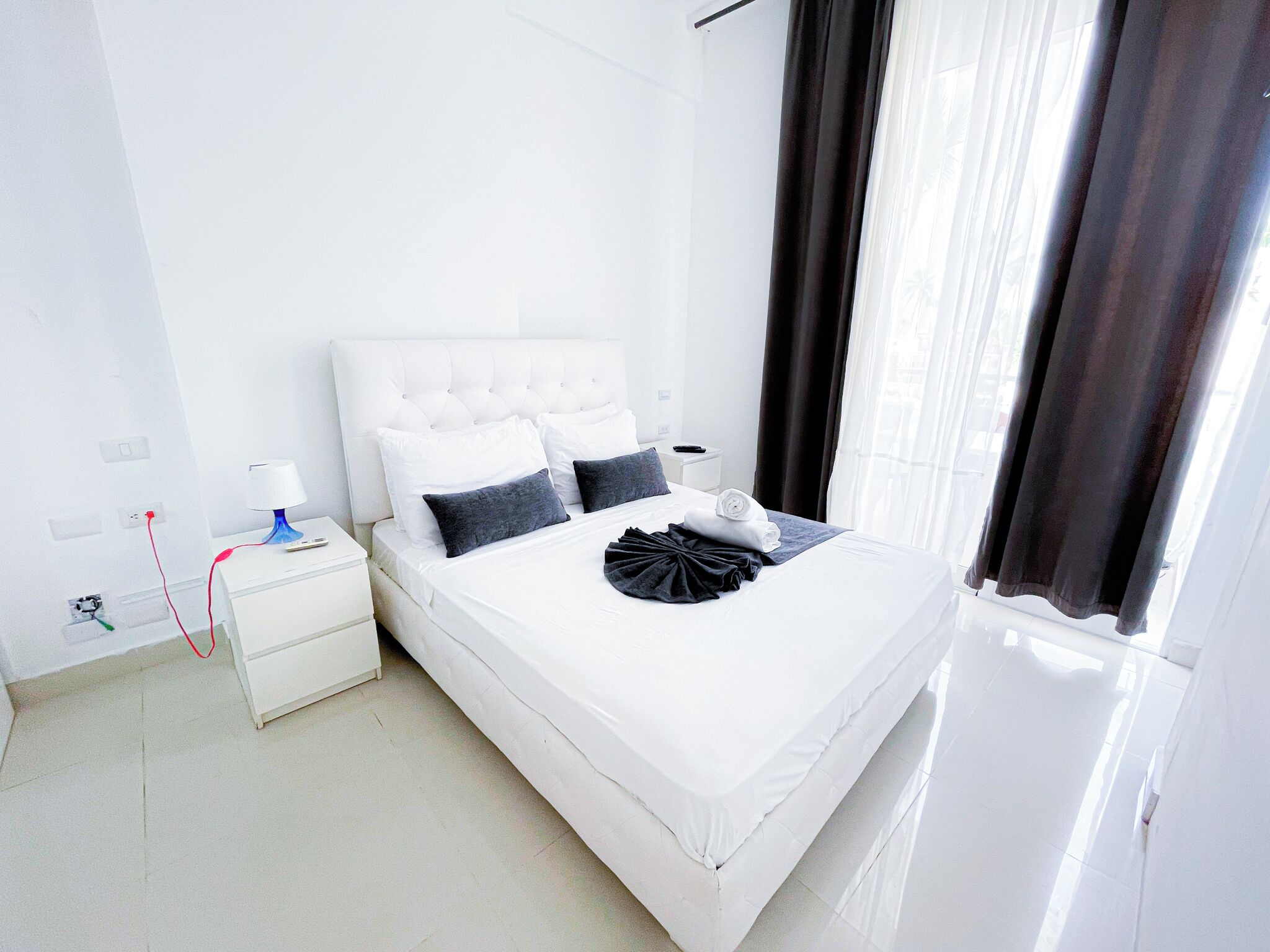 Habitación de hotel blanca con una cama grande, una mesita de noche, una lámpara y una ventana con cortinas.