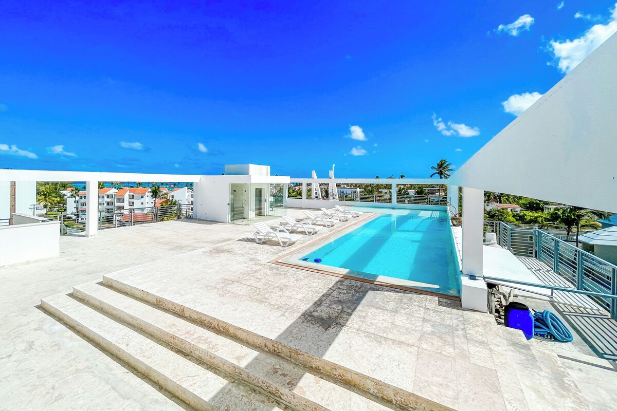 Uma piscina no terraço com água azul cristalina, rodeada por degraus e um edifício branco.