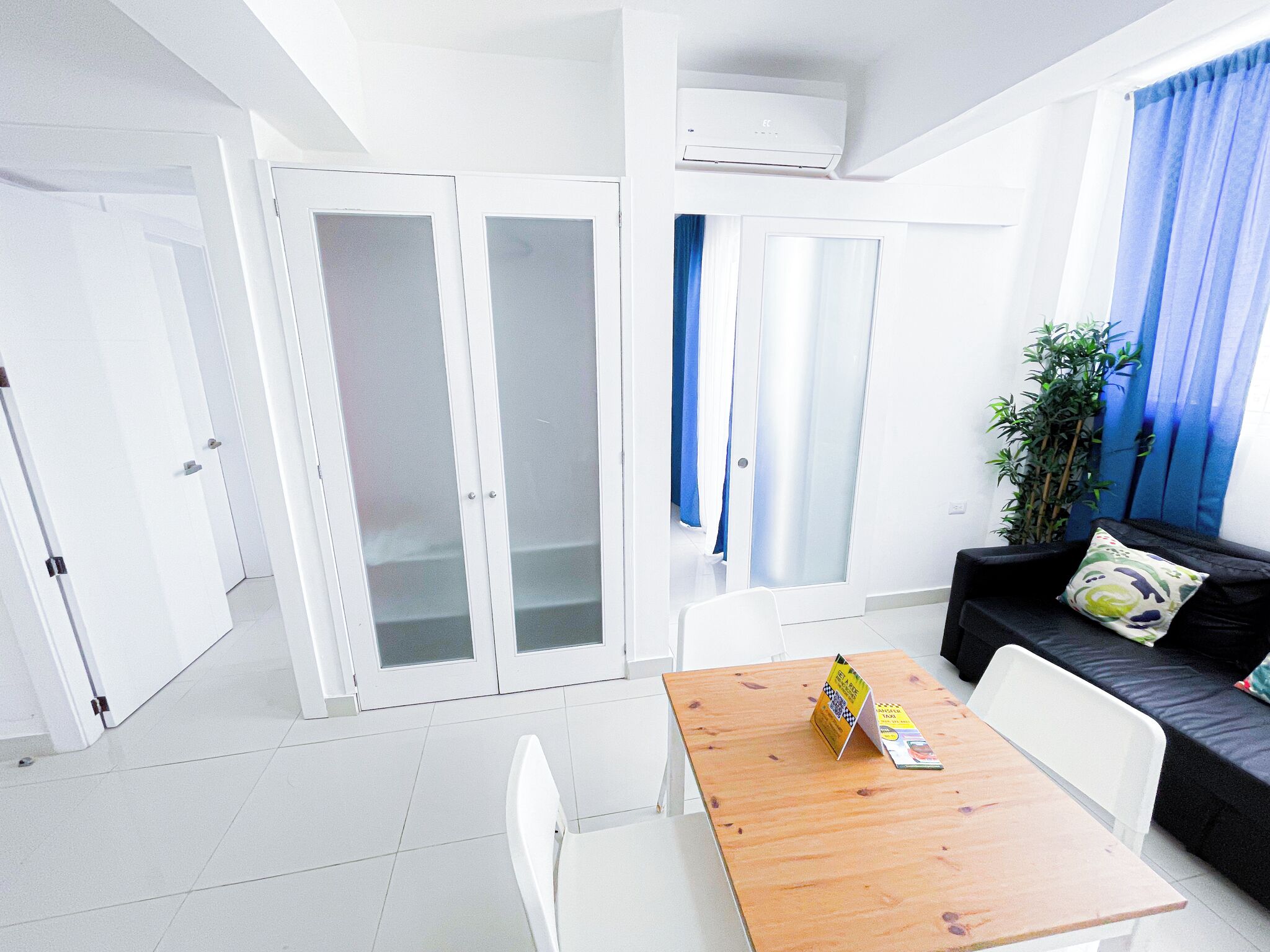 Een moderne, goed verlichte kamer met een houten tafel, witte stoelen, een zwarte bank met kussen, een potplant en een schuifdeur van glas.