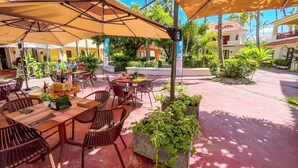 Ein Straßencafé mit Tischen und Stühlen unter großen Sonnenschirmen, umgeben von Grünpflanzen und Palmen.