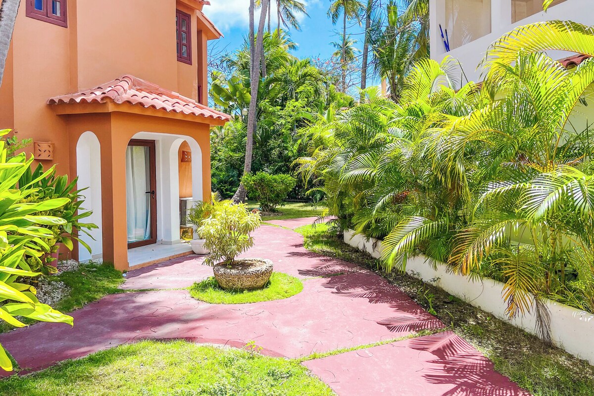 Uma trilha leva a um edifício com telhado de telhas vermelhas e uma pequena área de jardim com palmeiras e um vaso de planta.