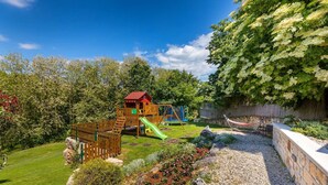 A playground with a slide and climbing structures, surrounded by a stone wall and lush greenery.
