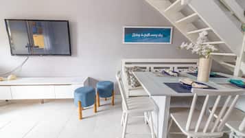 Een moderne eethoek met een witte tafel, blauwe stoelen en een witte bank. Er is een televisie aan de muur bevestigd en een ingelijste quote boven de tafel.