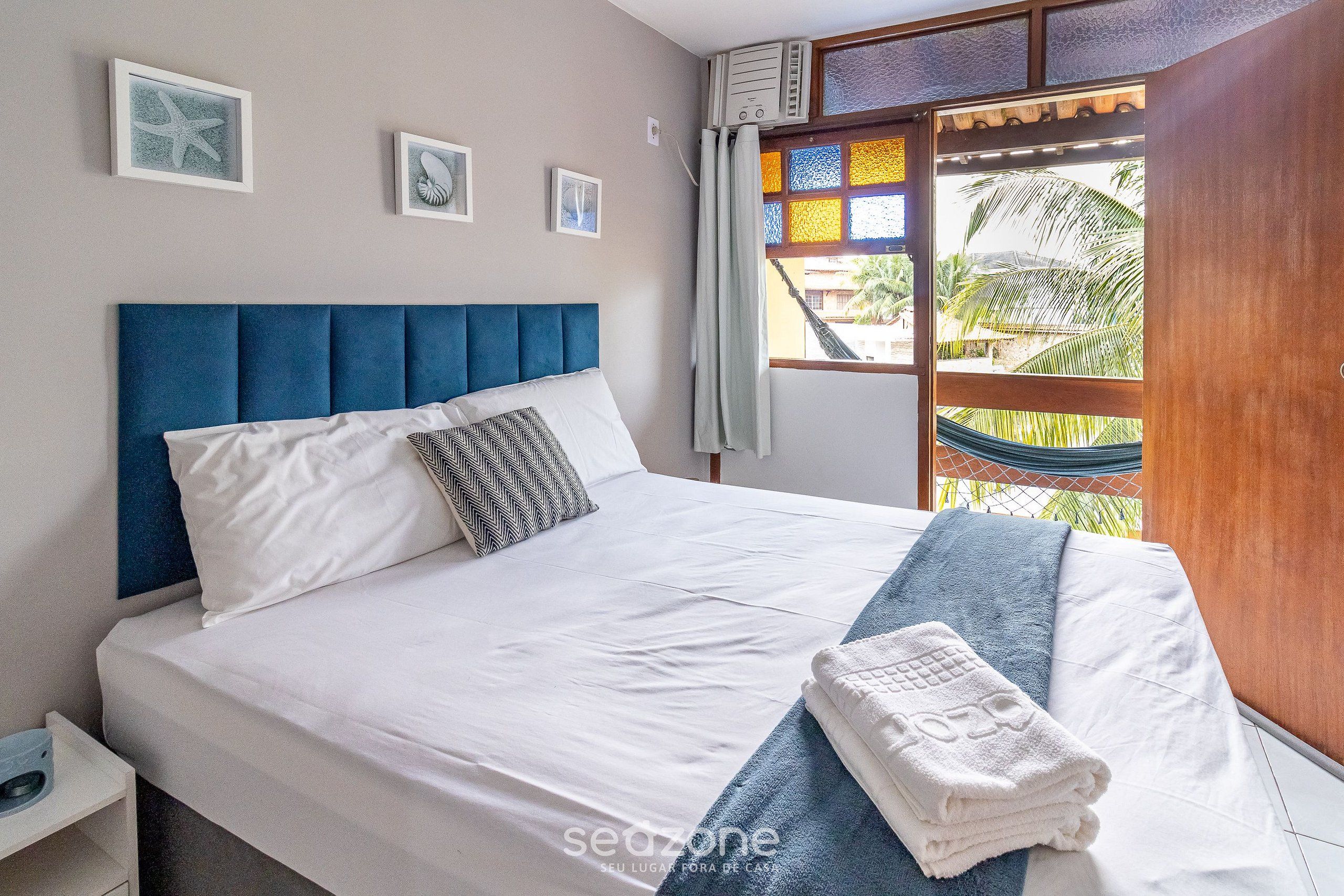 Uma cama bem arrumada com roupa de cama branca e azul, cabeceira azul e uma rede visível pela janela.