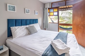 Uma cama bem arrumada com roupa de cama branca e azul, cabeceira azul e uma rede visível pela janela.
