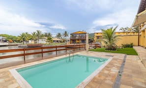 A swimming pool with a wooden deck, surrounded by palm trees and buildings with yellow and brown facades.