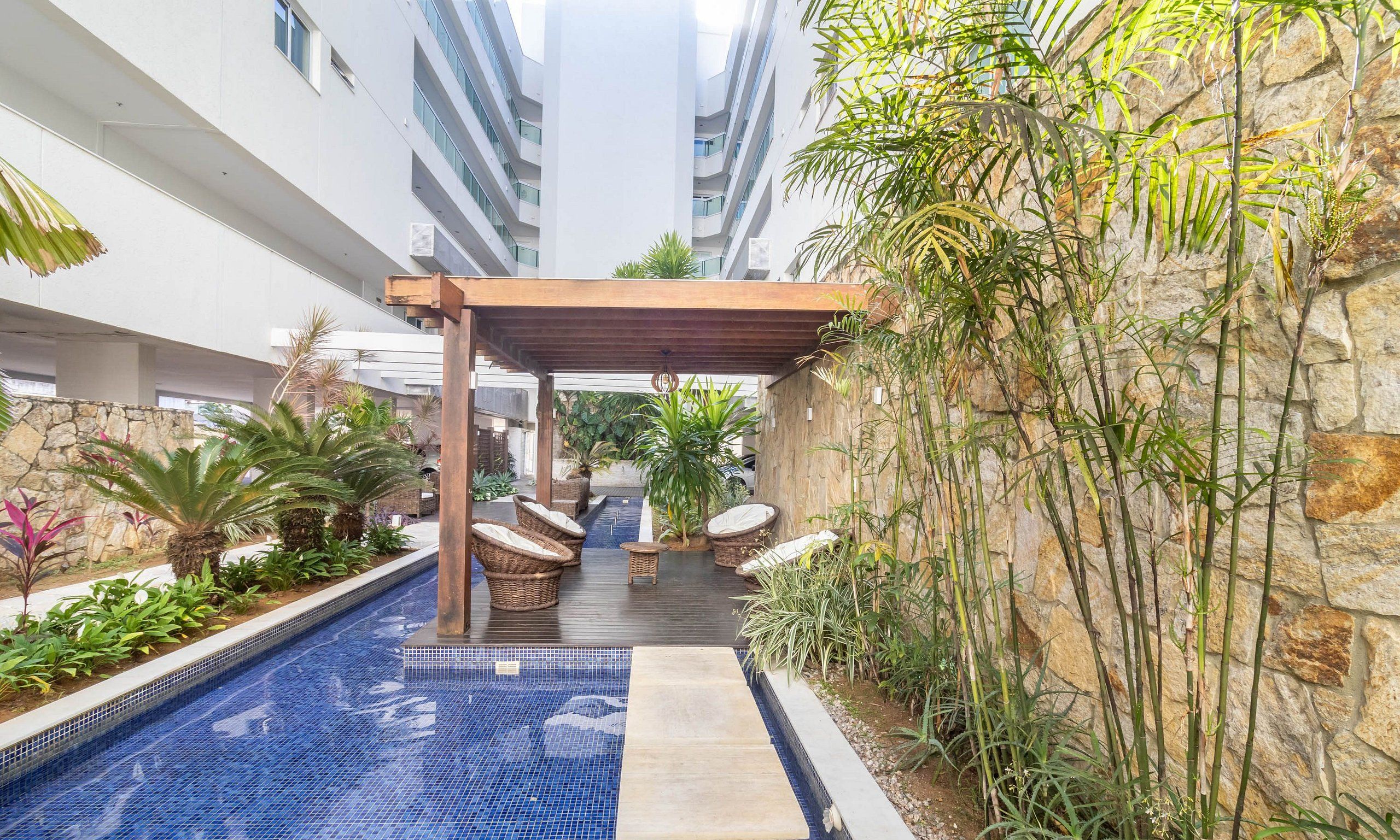 Uma área de piscina moderna ao ar livre, com pérgula, espreguiçadeiras e diversas plantas.