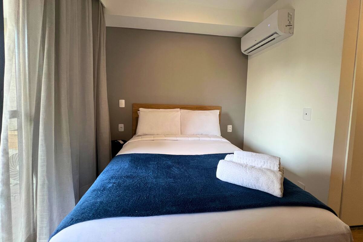 A hotel room with a bed, a window with curtains, an air conditioning unit, and a wall socket.
