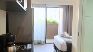 Habitación de hotel moderna con kitchenette, una cama con toallas enrolladas y vistas a la vegetación por la ventana.
