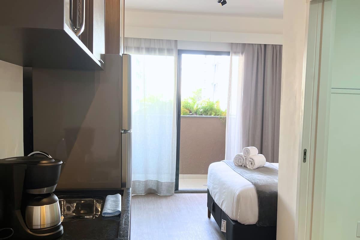 A modern hotel room with a kitchenette, a bed with rolled towels, and a view of greenery through the window.