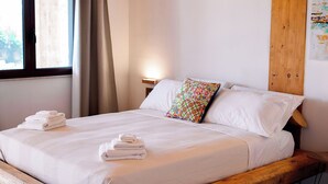 Uma cama de madeira com lençóis brancos e um travesseiro colorido em um quarto com teto de madeira e janela com cortinas.