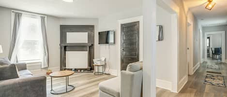 A modern living room with a sofa, armchair, and coffee table. There is a flat-screen TV mounted on the wall and a fireplace. The room has hardwood flooring and a large window with curtains.