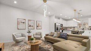 Un salon moderne avec un canapé beige, deux fauteuils, une table basse et un tapis. Le mur est décoré de trois tableaux abstraits. On y trouve également un coin cuisine avec des meubles blancs et des appareils en acier inoxydable, ainsi qu’un luminaire suspendu.