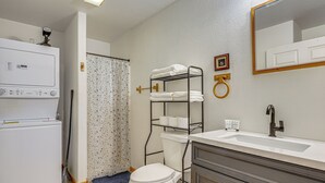 Un baño con inodoro blanco, lavamanos con espejo, toallero y cortina de ducha.
