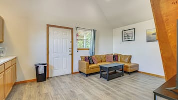 Um quarto com piso de madeira, um sofá bege com almofadas, uma mesinha, uma porta, uma janela com cortinas e um quadro fixado na parede.