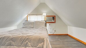 Un dormitorio con techo inclinado, una cama con cubrecama acolchado, un aire acondicionado de pared y una ventana con cortinas.