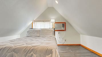 Een slaapkamer met een schuine plafond, een bed met een gewatteerde spreidekking, een luchtverkoeler aan de muur en een raam met gordijnen.