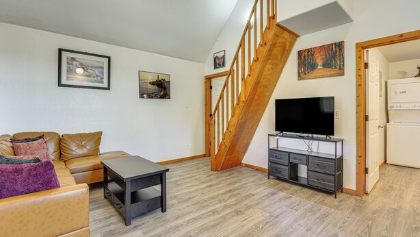 Una sala de estar con un sofá, una mesa de centro, un televisor sobre un mueble, una escalera y una puerta que da a otra habitación.