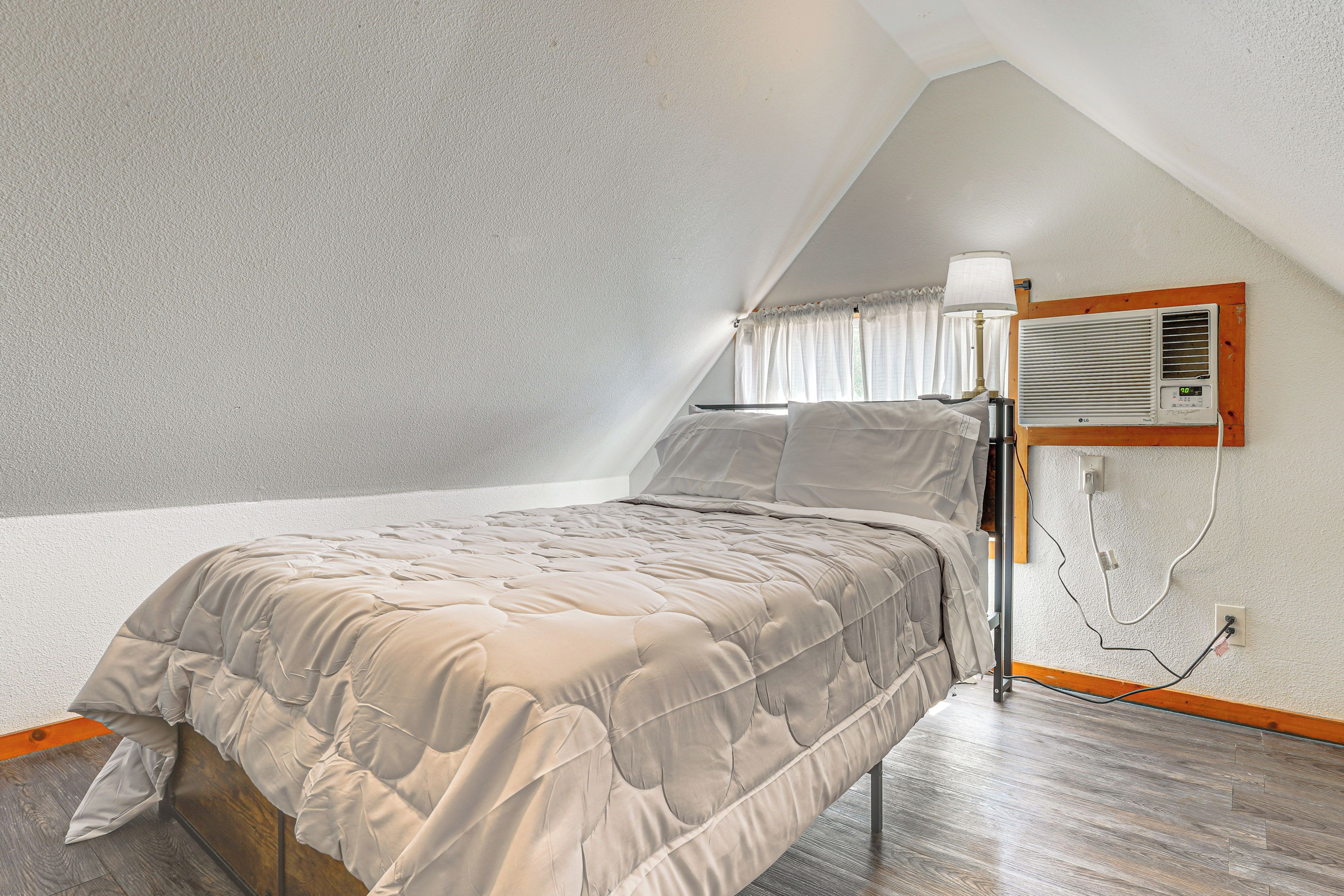 A bedroom with a sloped ceiling, a bed with a beige quilted bedspread, a bedside table with a lamp, and an air conditioning unit mounted on the wall.