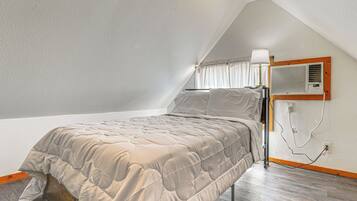 A bedroom with a sloped ceiling, a bed with a beige quilted bedspread, a bedside table with a lamp, and an air conditioning unit mounted on the wall.