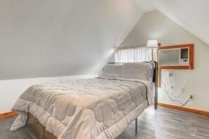 Een slaapkamer met een schuine kap, een bed met een beige gewatteerde spreideken, een nachtkastje met een lamp en een airconditioner aan de muur bevestigd.