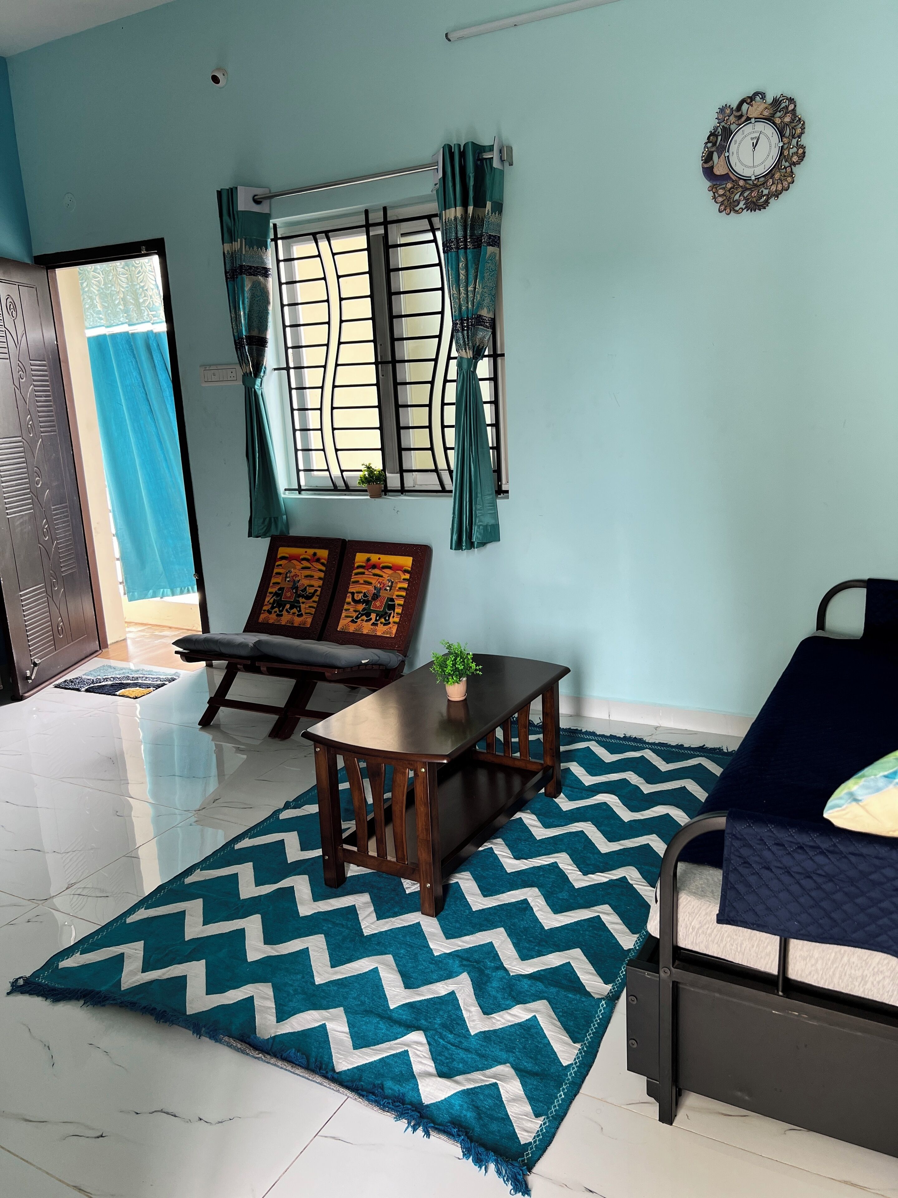 A room with a bed, a table with a plant, a sofa, a window with curtains, and a wall clock.