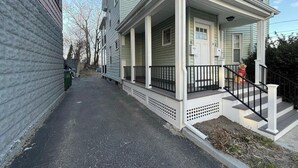 A two-story residential building with a front porch and a small front yard. - Cozy 4BR Home by Tufts w/ Subway Access to Downtown, MIT, Harvard + Free Parking (Medford)
