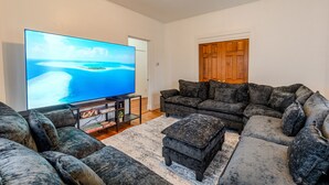 A living room with a large flat-screen TV, a dark-colored sectional sofa, a matching ottoman, and a wooden door. - Cozy 4BR Home by Tufts w/ Subway Access to Downtown, MIT, Harvard + Free Parking (Medford)