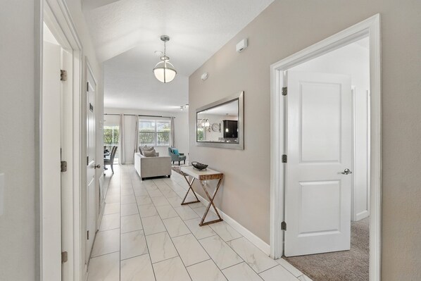 A hallway with white tiled floors, a ceiling light, a white door, and a small table with a bowl.
