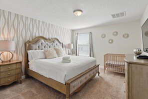 A bedroom with a large bed, a wooden nightstand, a bedside table, a crib, and a dresser.