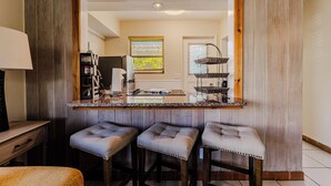 Una cocina con barra de madera, tres taburetes acolchados, un espejo grande y una ventana con persianas.