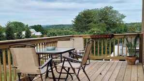 A wooden deck with a table and chairs, surrounded by a wooden railing, overlooking a lush green landscape. - Magnolia House (Broadway)