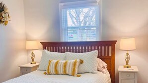 A neatly made bed with white bedding and striped pillows, flanked by two bedside tables with lamps, in a room with a window and a view of trees. - Magnolia House (Broadway)