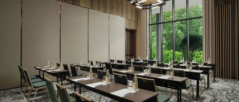 A conference room with multiple tables and chairs, large windows with a view of greenery, and a patterned ceiling.