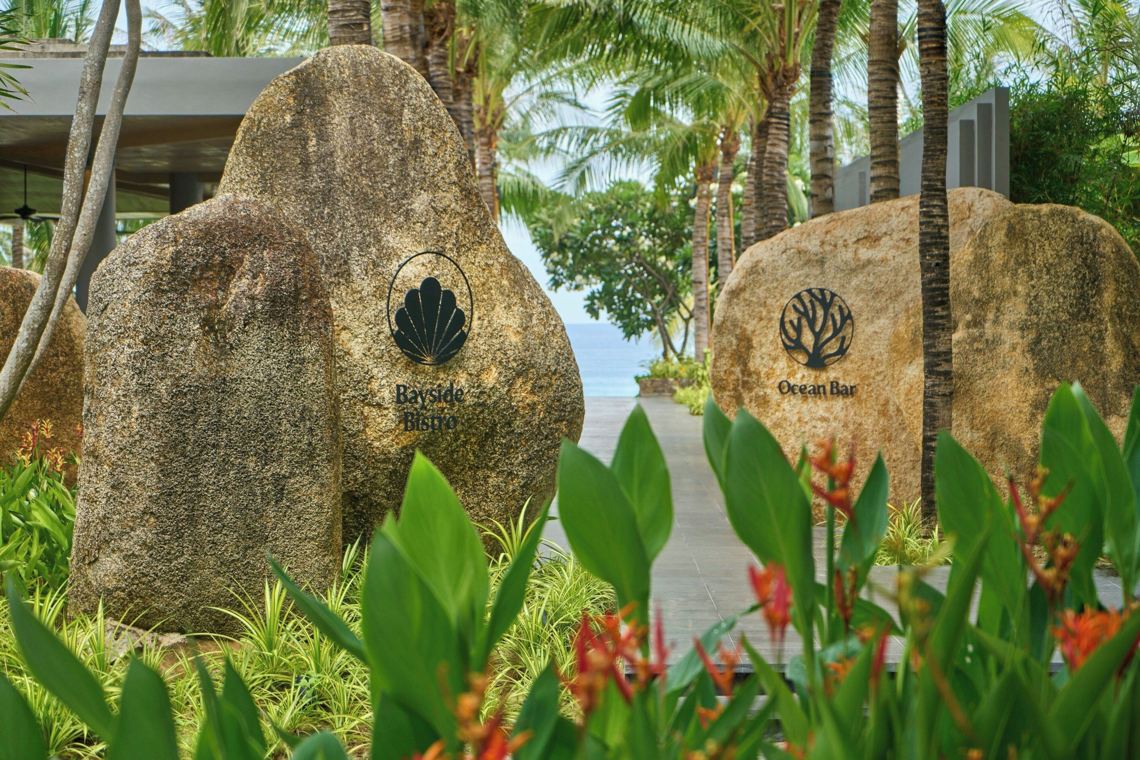 Two large rocks with logos and names, surrounded by greenery and palm trees.