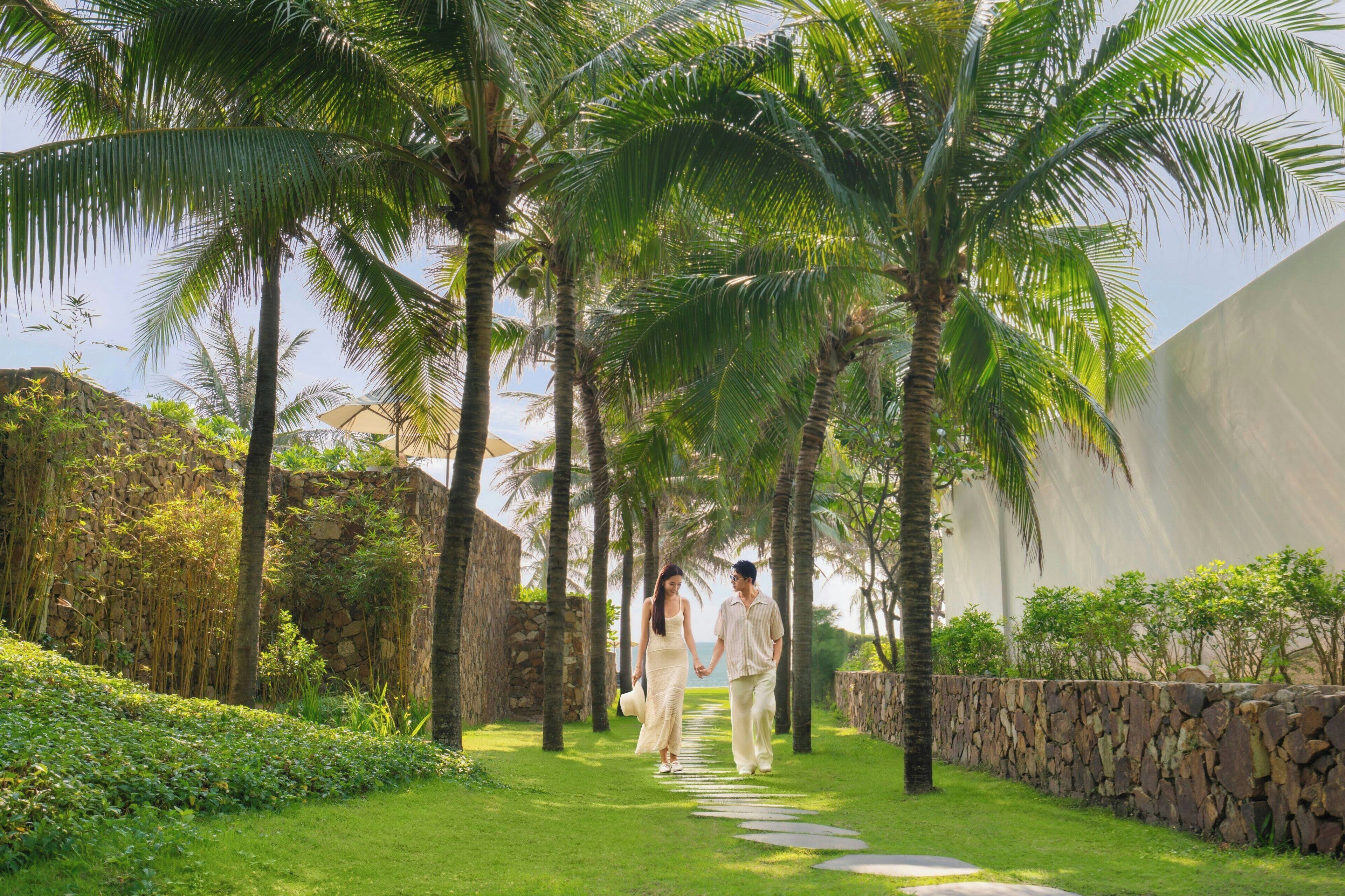 A pathway lined with palm trees and a stone wall, leading to a building with a stone facade.