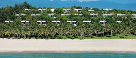 Um resort com várias acomodações em estilo de bangalô, uma praia e montanhas ao fundo.