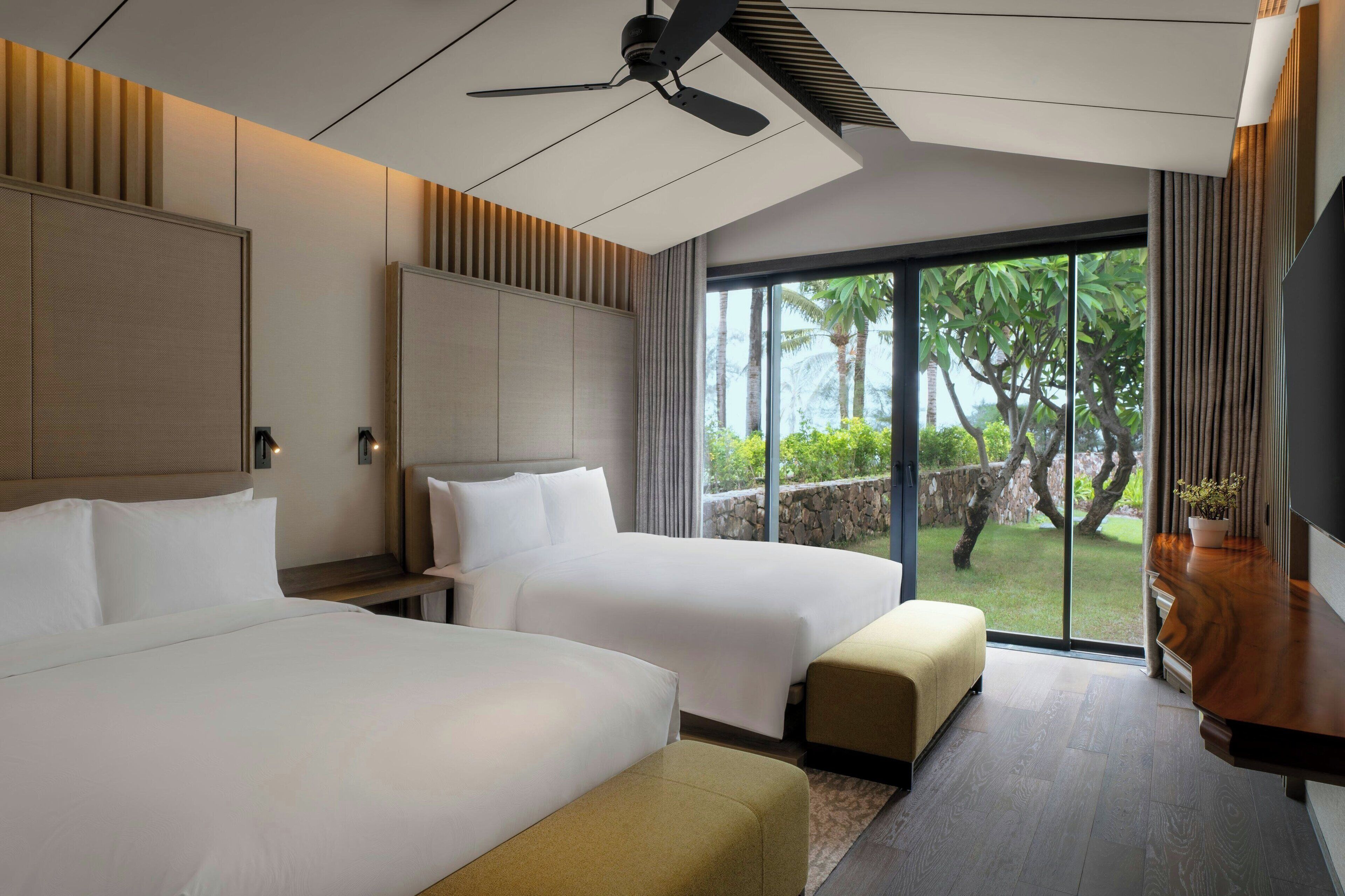 A hotel room with two beds, a ceiling fan, a large window with a view of greenery, and a wooden side table with a plant.