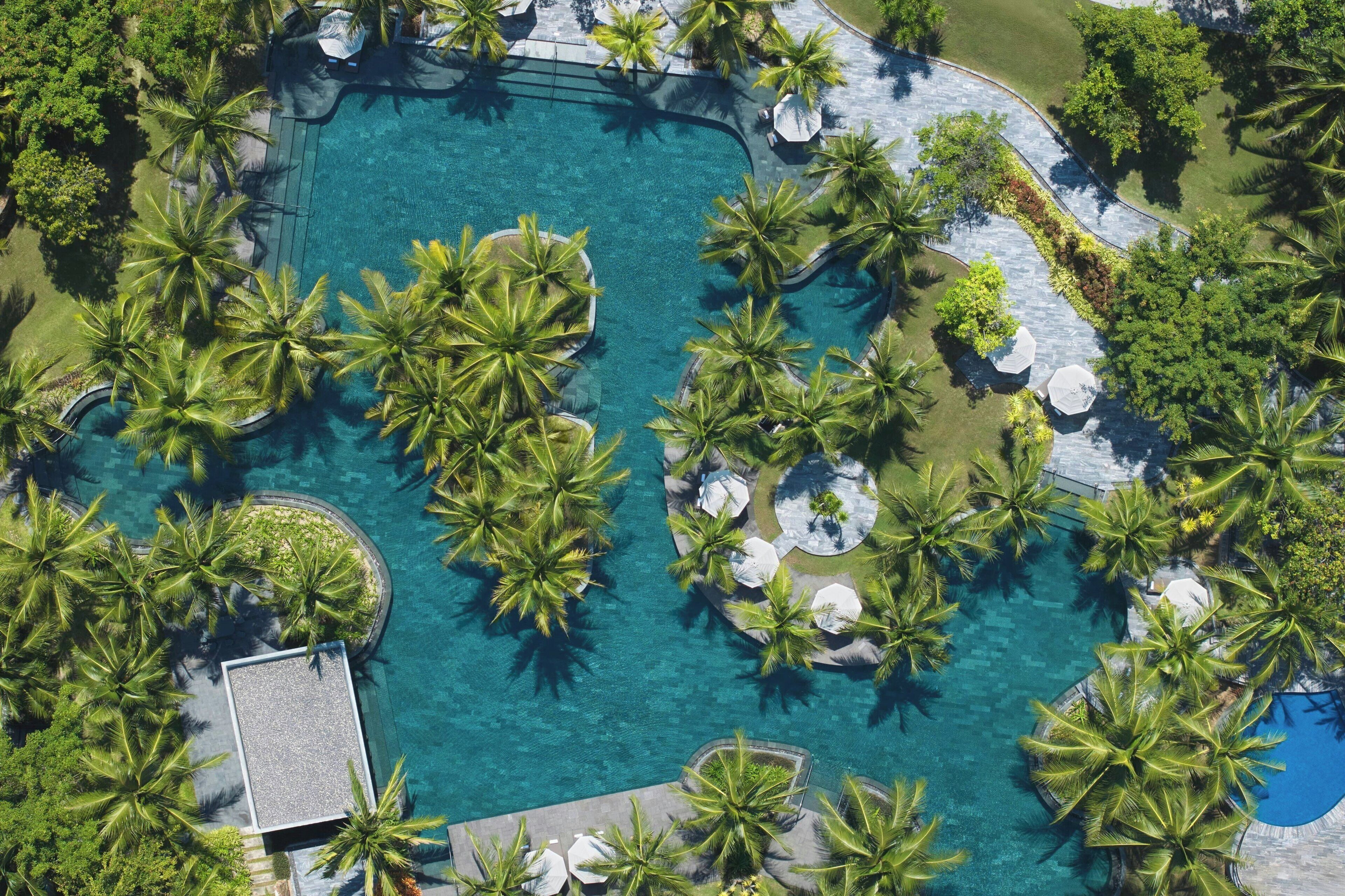 Luchtbeeld van een resort met een groot centraal zwembad, omgeven door palmbomen en schaduwrijke plekken.