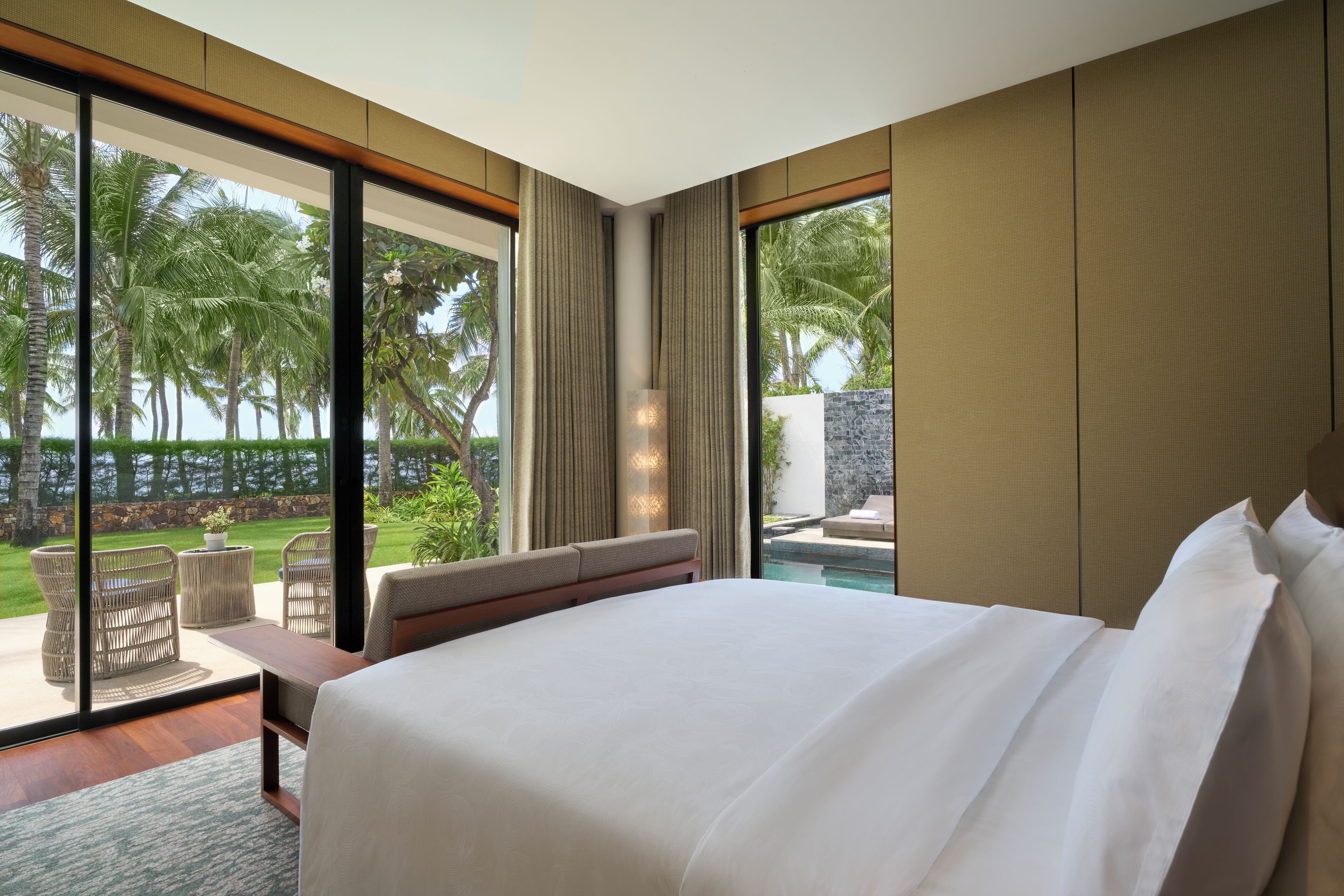 Un dormitorio con una cama grande, un área de descanso con vista a una piscina y palmeras, y un ventanal amplio.