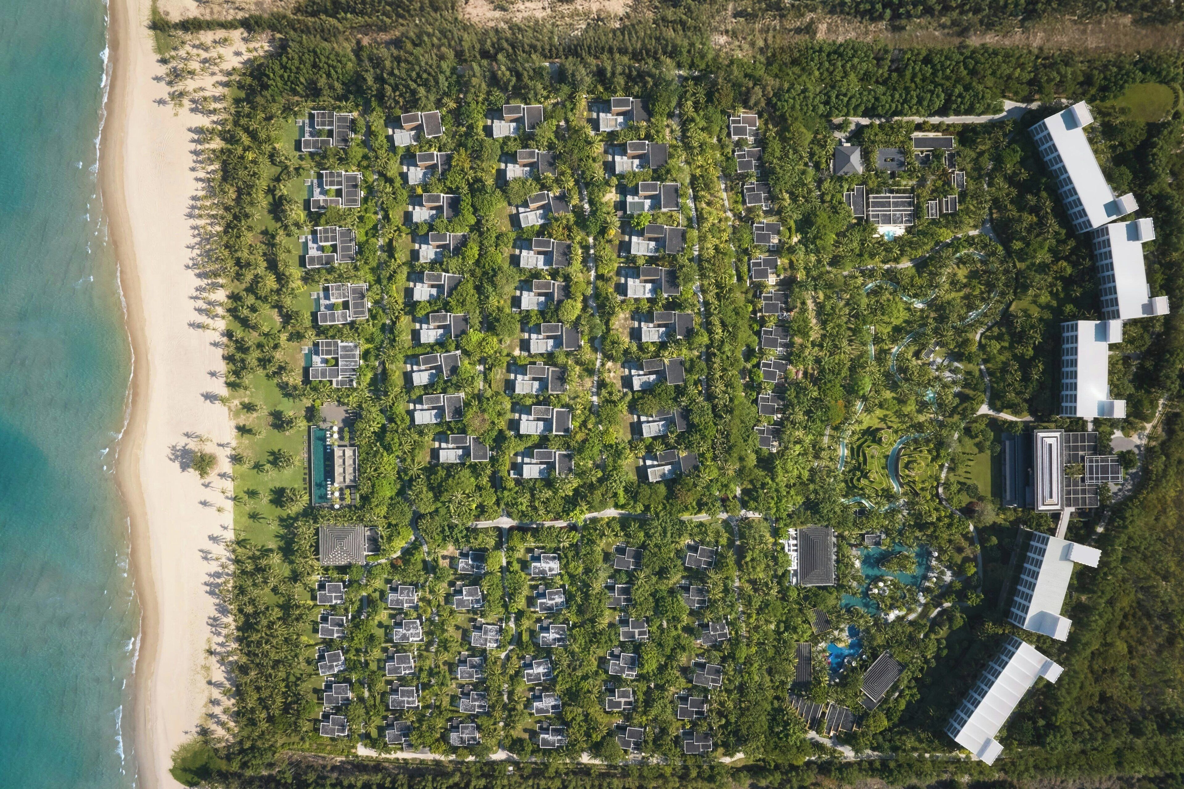 Vista aérea de un complejo turístico con varias cabañas dispuestas en filas, una playa y una zona de piscina.