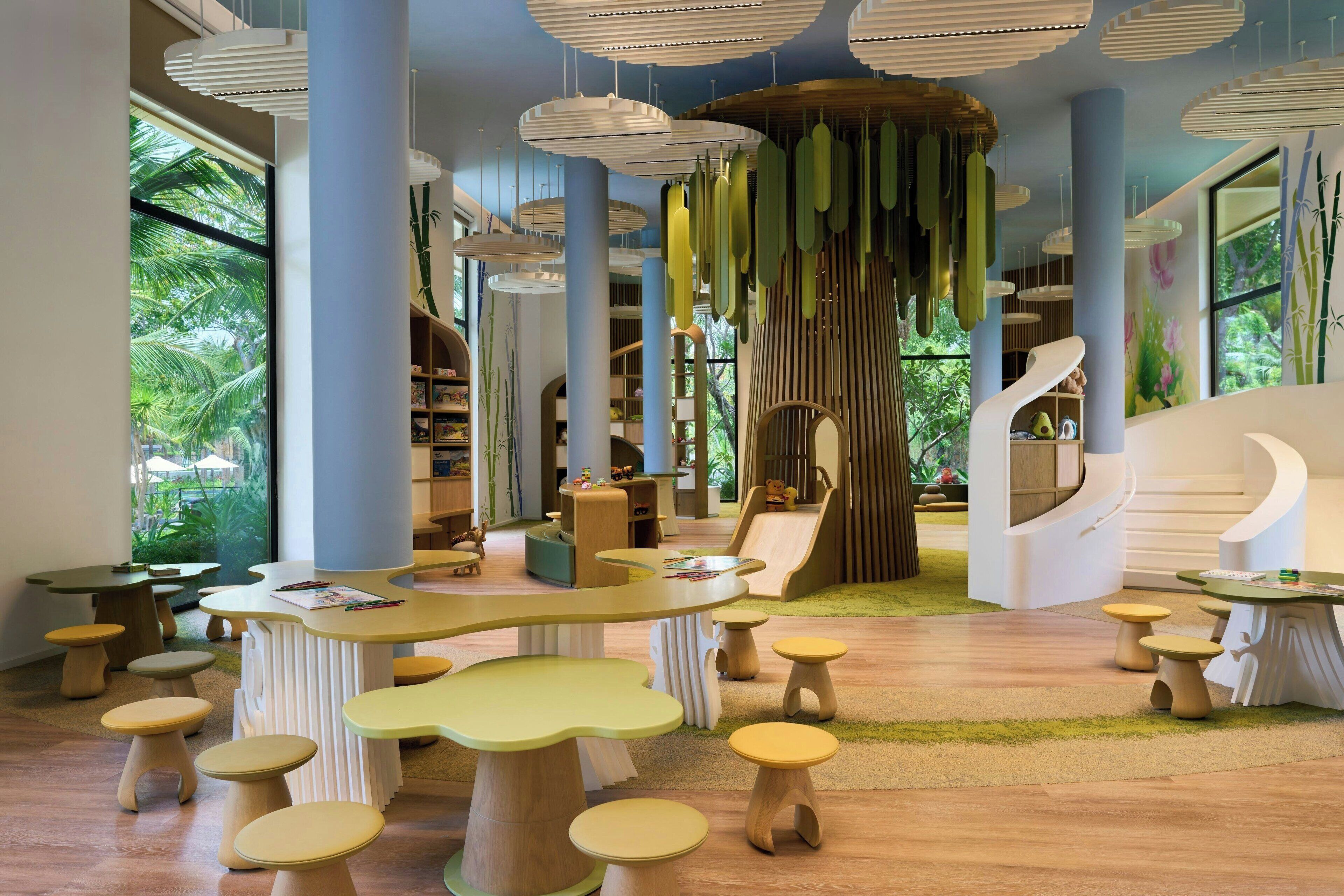 Un espacio interior moderno con mobiliario único, que incluye mesas y taburetes curvos, y una zona de juegos central con toboganes y estructuras para escalar.