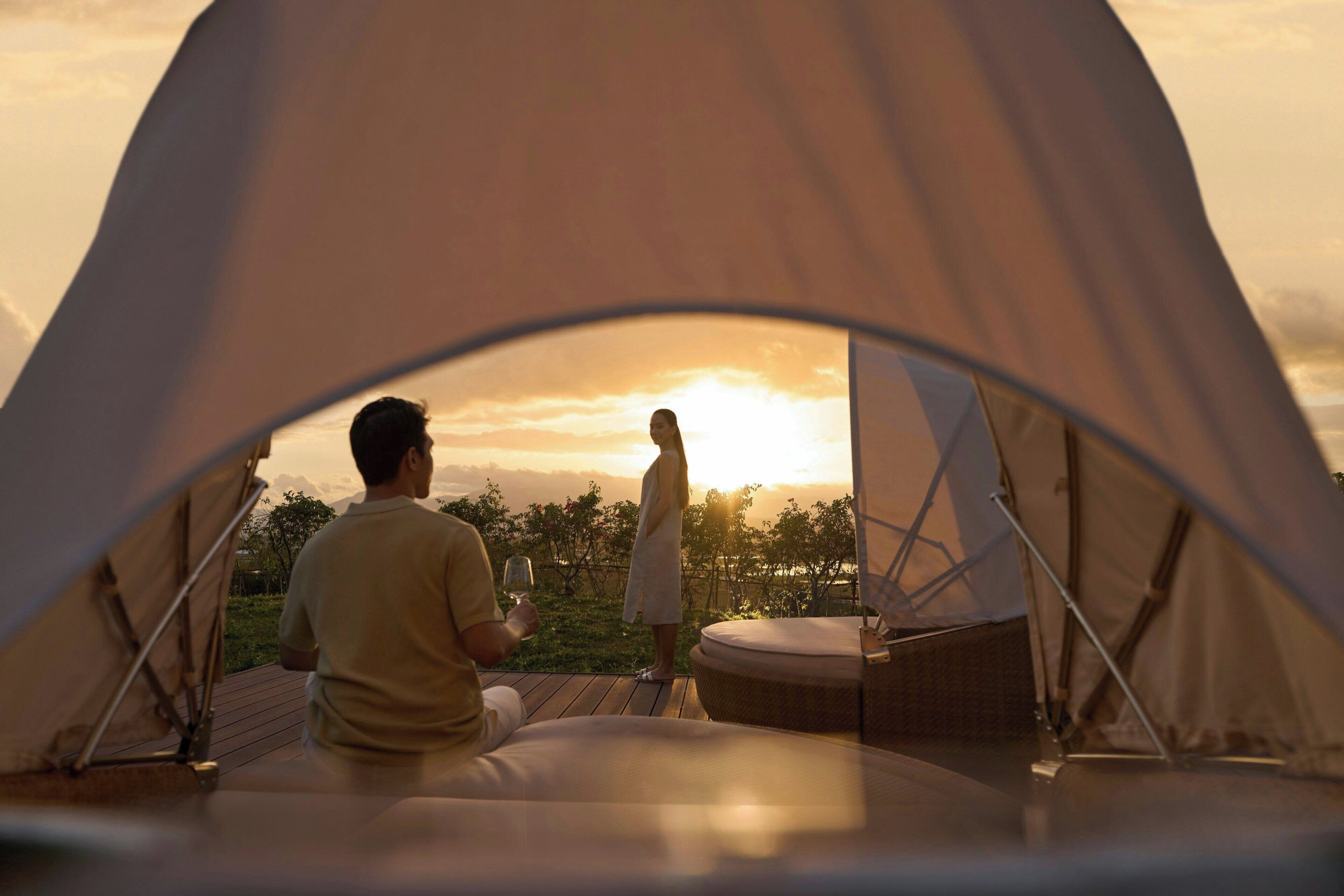 A tent with a view of a sunset, a person standing, and another person sitting inside.