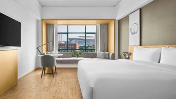 A hotel room with a large bed, a desk, a chair, a TV, and a window with a city view.