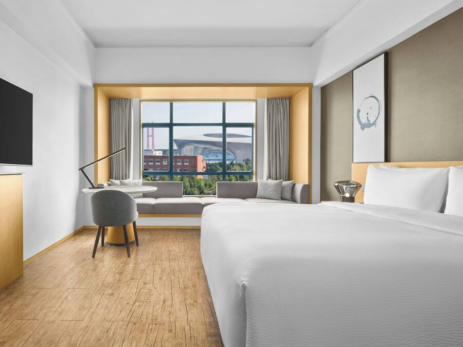 A hotel room with a large bed, a desk, a chair, a TV, and a window with a city view.