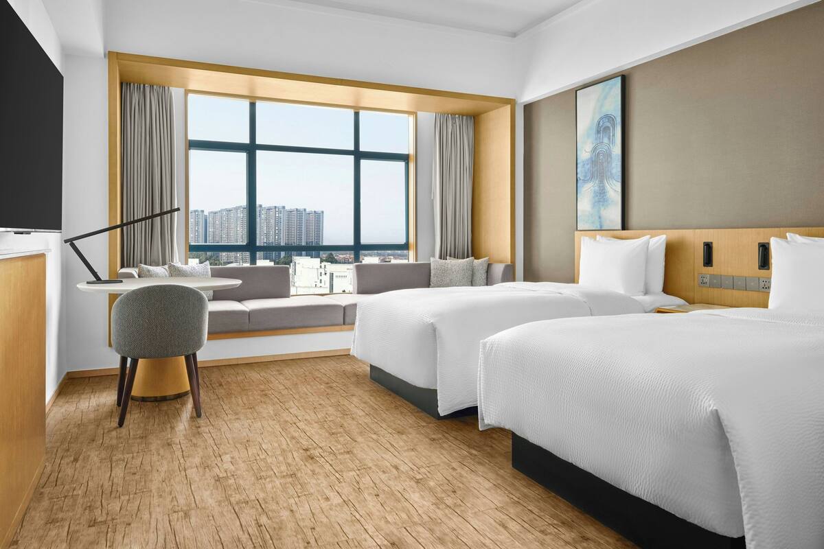 Hotelkamer met groot raam, een bank, twee bedden, een bureau en een stoel.