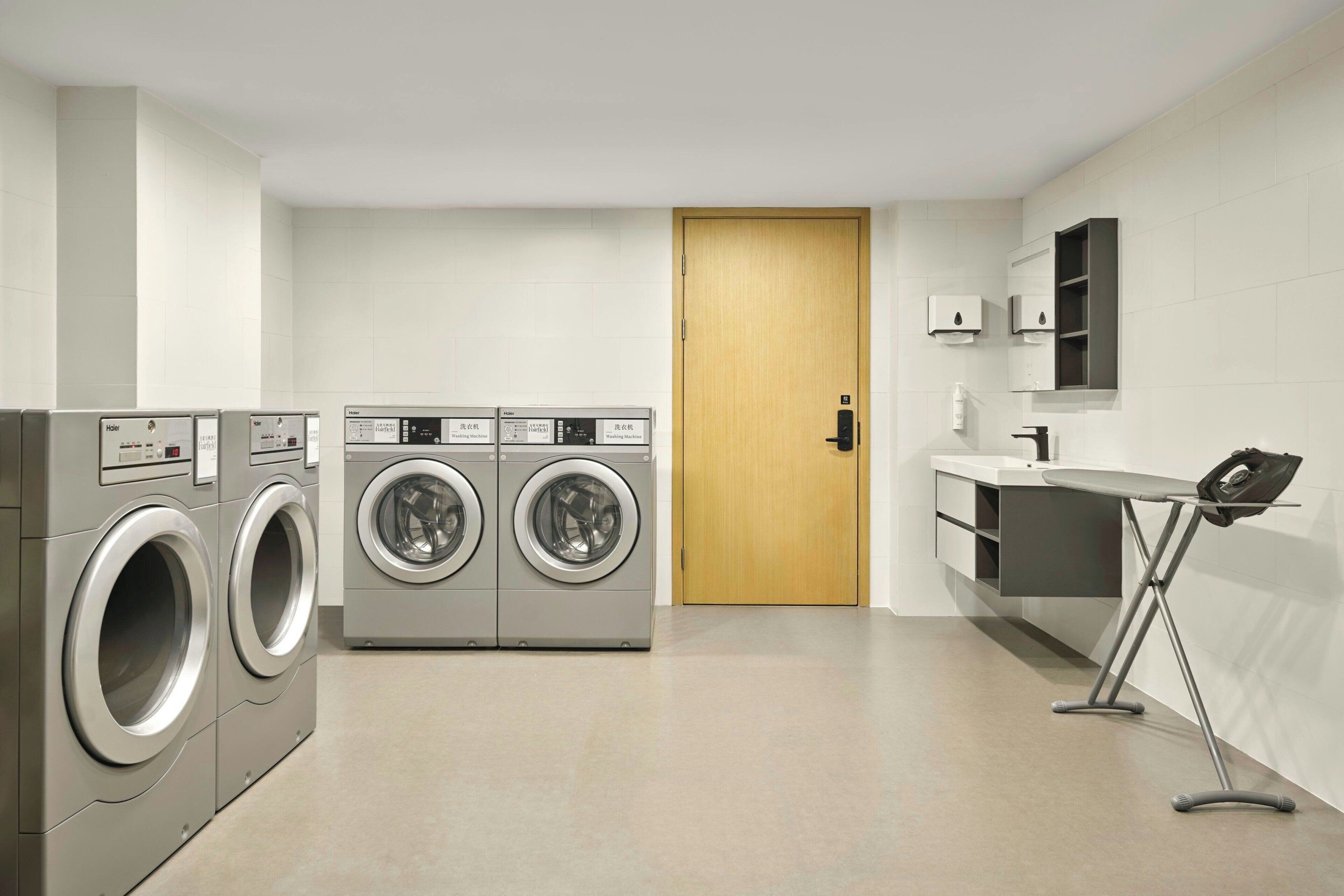 A modern laundry room with two front-loading washing machines, a wooden door, and a small ironing station.