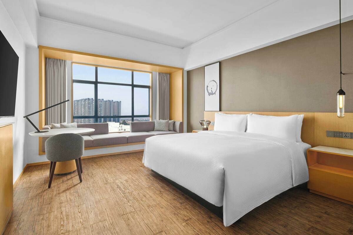 Hotelkamer met een groot bed, een zithoek met een bank en fauteuils, een eettafel en een groot raam met uitzicht op de stad.