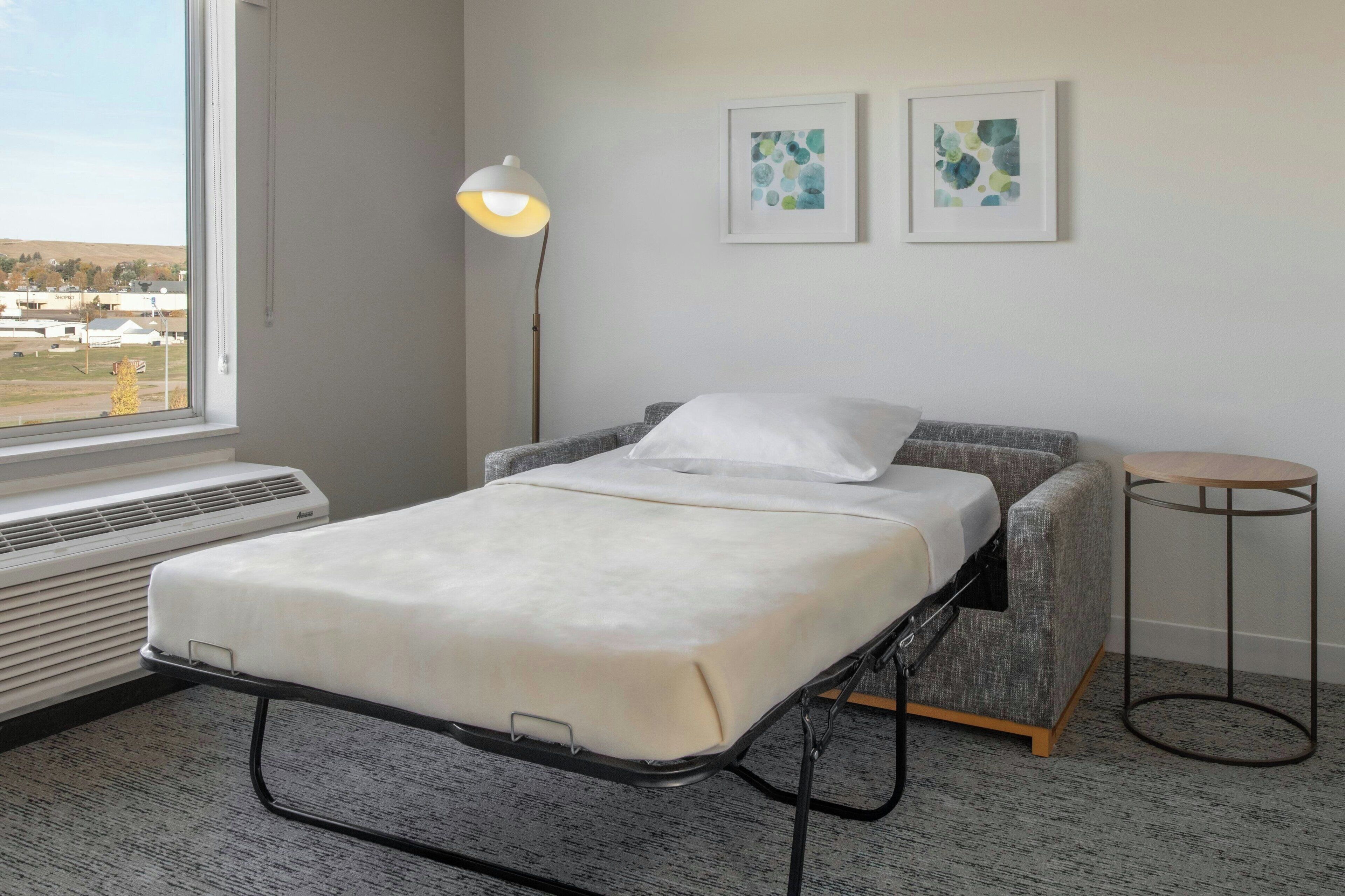 A single bed with a mattress and pillows, a bedside table, a floor lamp, two framed artworks, and a window with a view of buildings and trees.