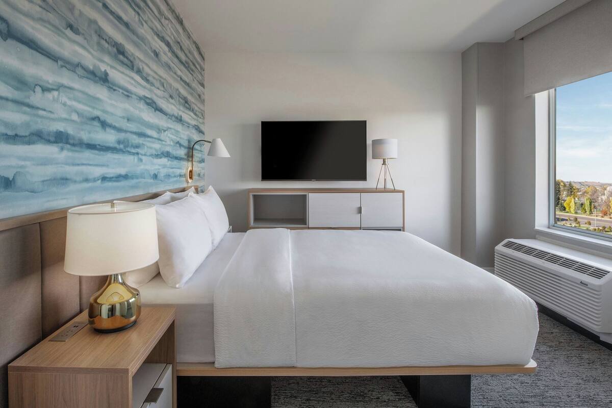 Une chambre d’hôtel moderne dotée d’un grand lit, d’un téléviseur à écran plat, d’une lampe de chevet et offrant une vue sur la ville par la fenêtre.